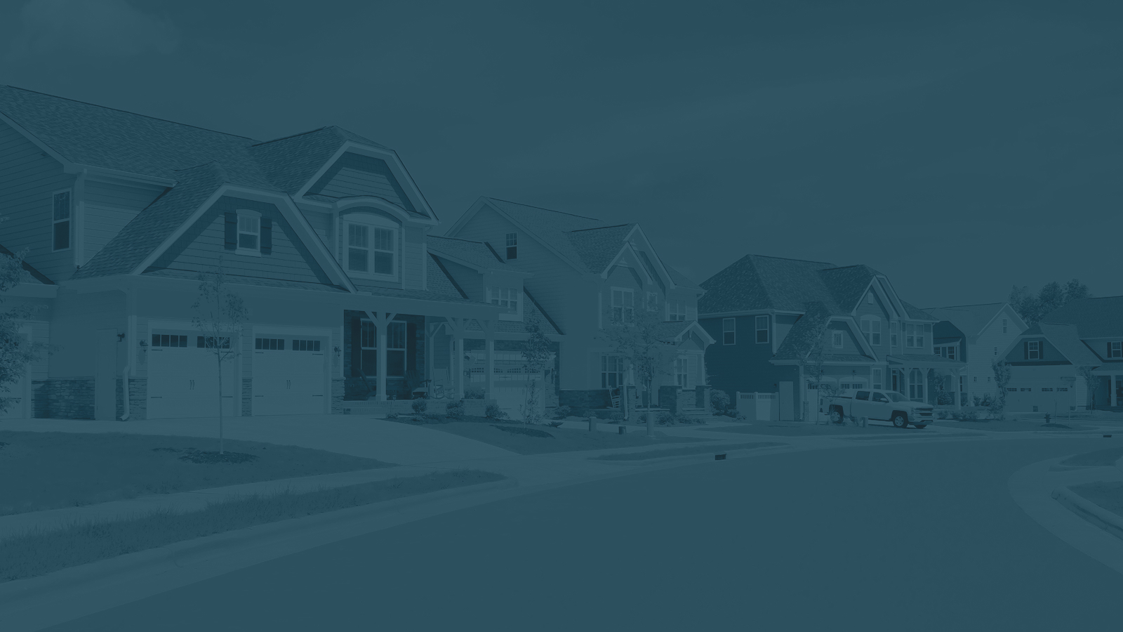 A suburban street lined with modern houses featuring various architectural styles. The homes have well-maintained lawns, driveways, and some have porches. A white pickup truck is parked in front of one of the houses. The sky is partly cloudy, adding a bright atmosphere to the neighborhood scene.