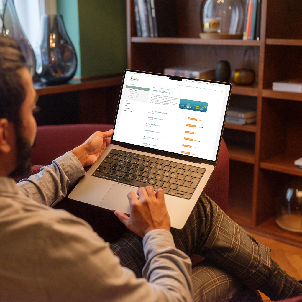 A person is seated on a couch, using a laptop. The laptop screen displays a webpage titled "Home Sales Reports," featuring a list of reports with orange buttons for access. The individual is wearing a light blue shirt and has a beard. The background includes a bookshelf with various items and a green wall. The setting appears cozy and well-lit.