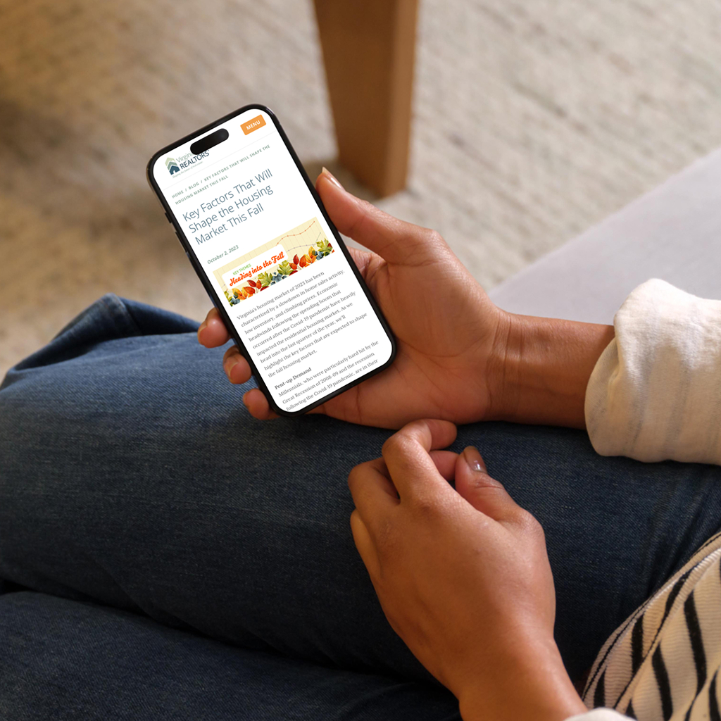A person is sitting on a couch, holding a smartphone in their right hand. The phone displays an article titled "Key Factors That Will Shape the Housing Market This Fall," with a colorful header featuring fruits and vegetables. The person's left hand rests on their knee, and they are wearing a light-colored, long-sleeve shirt with a striped pattern. The background shows a soft, textured surface and a wooden table.