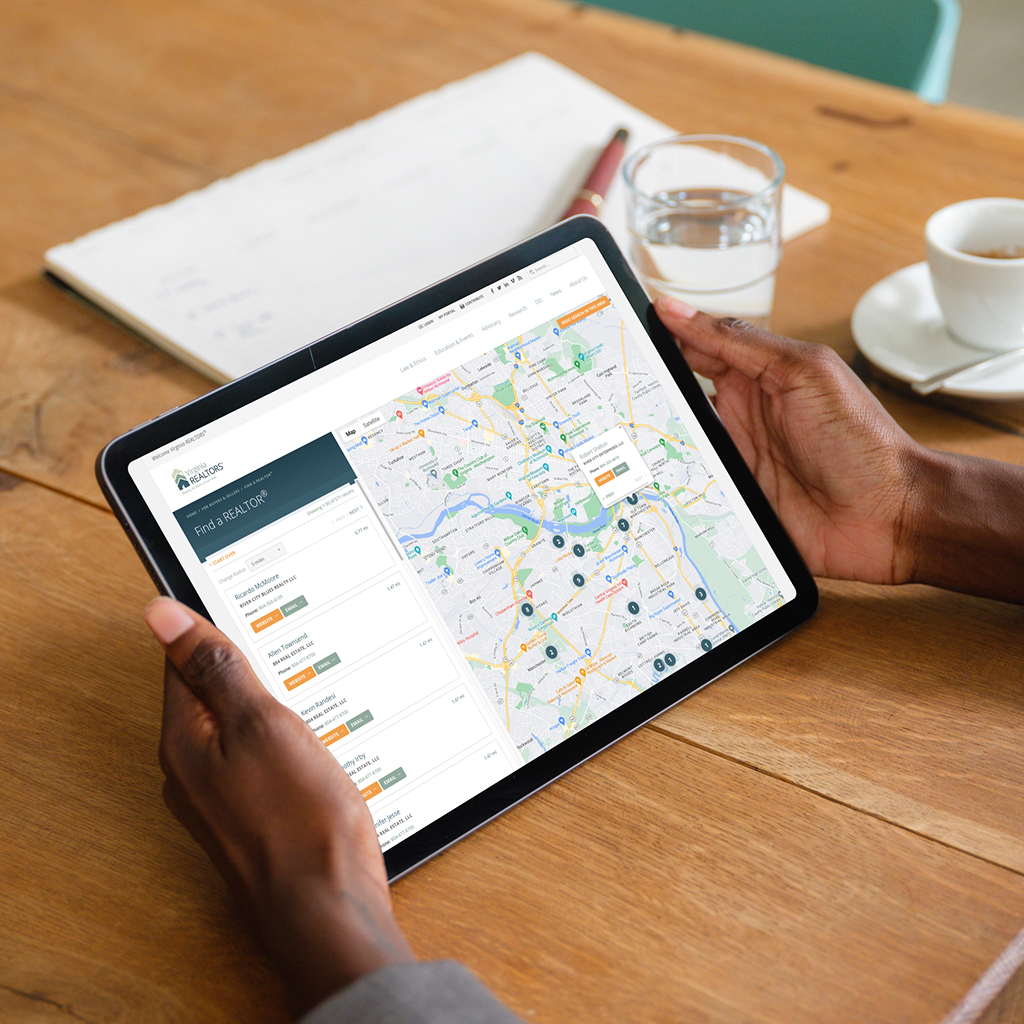 A person is holding a tablet displaying a map interface with various markers and listings on the left side. The map shows streets and locations, likely related to real estate or services. In the background, there is a wooden table with a glass of water, a cup of coffee on a saucer, and a notepad with a pen. The setting appears to be a casual workspace or café.