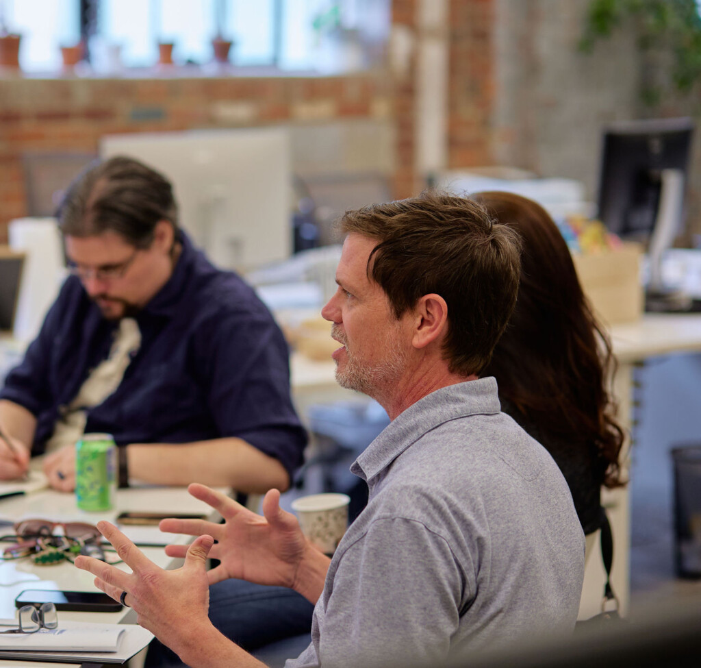 A group of people is engaged in a discussion in a modern office setting. In the foreground, a man with short hair and a light gray shirt is animatedly speaking, using hand gestures. Nearby, another man in a dark shirt is writing notes, while a woman with long hair is partially visible, listening attentively. A green soda can and several notebooks are on the table, along with a coffee cup and some colorful sticky notes. The background features brick walls and large windows with potted plants.
