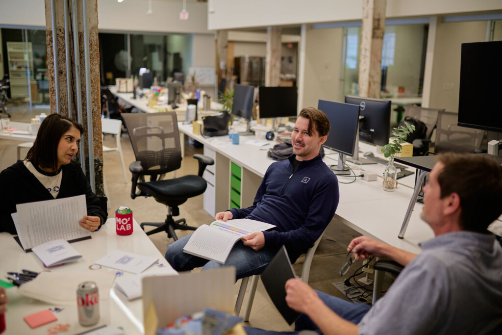 A group of three people is engaged in conversation in a modern office space. One person, a woman with dark hair, is sitting at a table, holding a document and looking attentively at another individual. The second person, a man with light brown hair, is seated across from her, smiling and also holding a document. The third person, a man with short hair, is partially visible on the right, holding a pair of glasses. The office features multiple desks with computers, plants, and various office supplies scattered around. The environment is bright and open, with large windows in the background.
