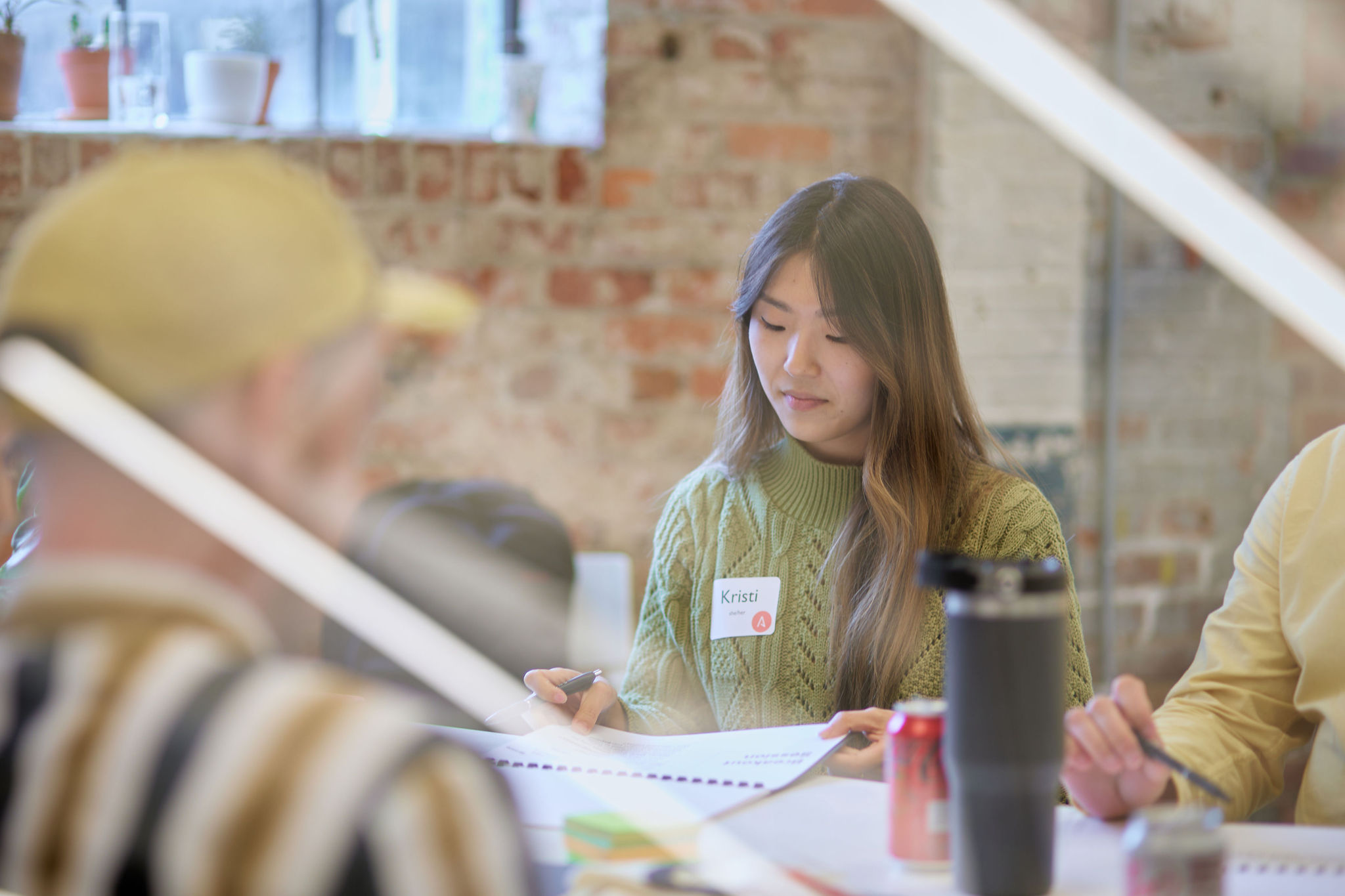 A strategist with long, wavy hair is seated at a table, focused on writing in a notebook. She wears a green knitted sweater and has a name tag on her chest. In the foreground, part of a person wearing a striped shirt is visible, along with a gray water bottle and a red soda can on the table.