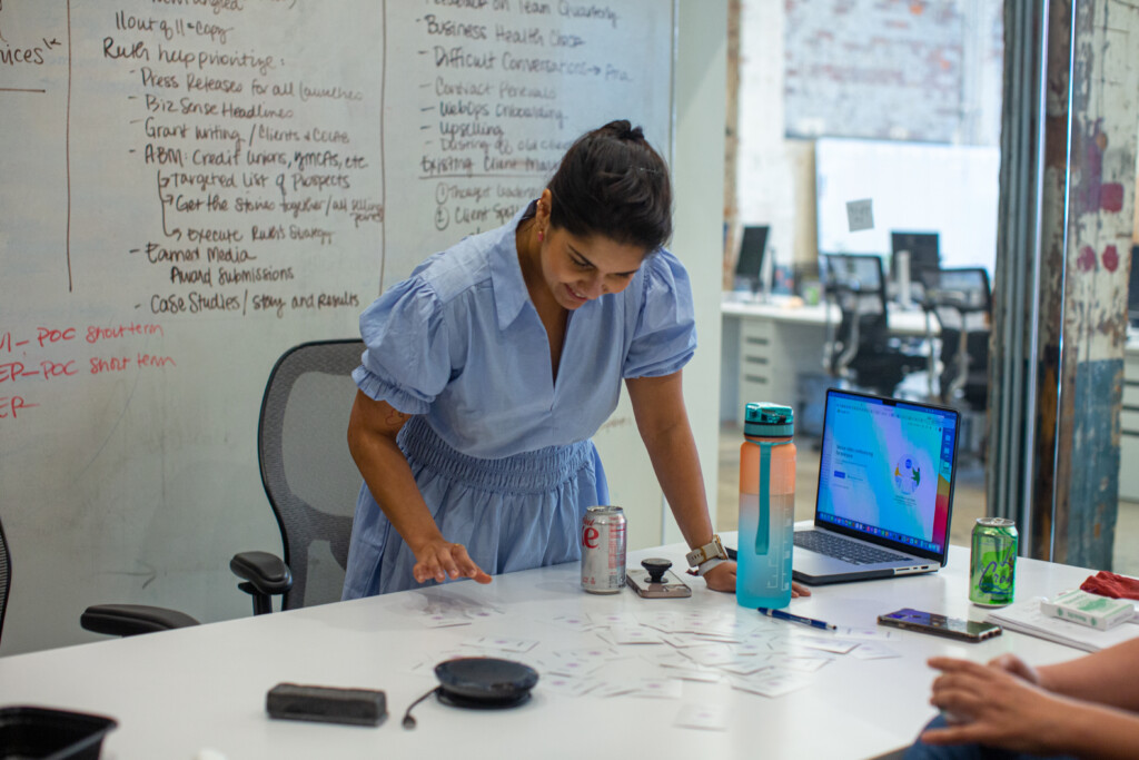 A woman in a light blue dress is leaning over a table, examining a spread of cards laid out in front of her. She has a can of Diet Coke and a smartphone nearby, along with a colorful water bottle. A laptop is open on the table, displaying a colorful screen. In the background, a whiteboard is filled with handwritten notes and lists, and office chairs are visible in a modern workspace setting.