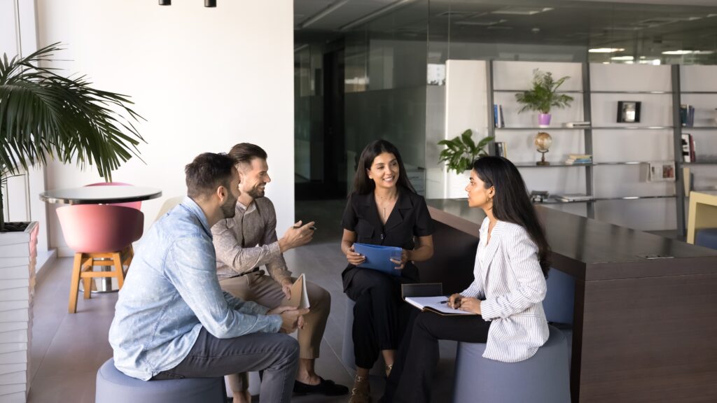 A group of five committee members engaged in a discussion in a modern office setting. They are seated on round stools, with two men and three women present. One woman, holding a tablet, appears to be leading the conversation, while the others listen attentively. The background features a large window with natural light, a potted plant, and a bookshelf filled with various items. The overall atmosphere is collaborative and professional.