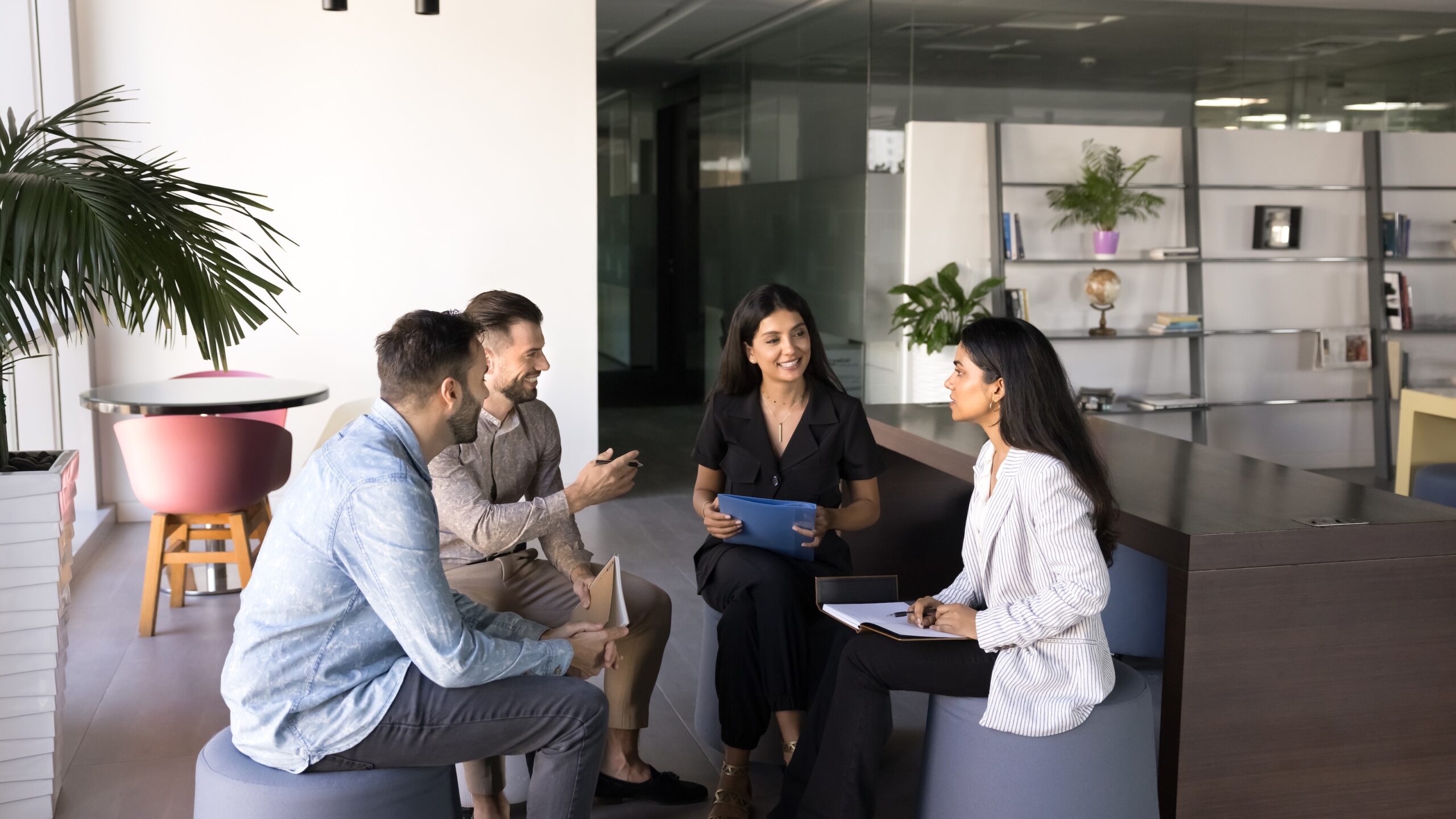 A group of five people is engaged in a discussion in a modern office setting. They are seated on round stools, with two men and three women present. One woman, holding a tablet, appears to be leading the conversation, while the others listen attentively. The background features a large window with natural light, a potted plant, and a bookshelf filled with various items. The overall atmosphere is collaborative and professional.