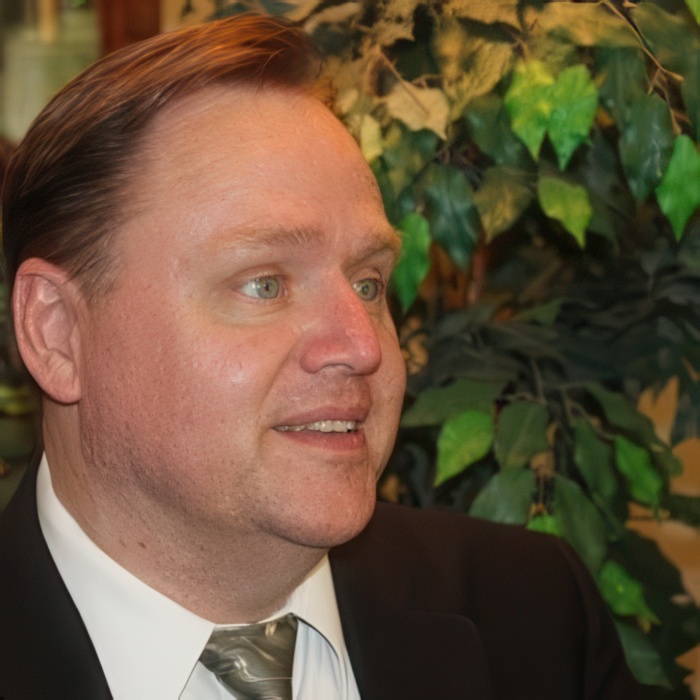 A man with short, slicked-back hair and light skin is shown in profile. He is wearing a black suit jacket over a white dress shirt and a light-colored tie. His expression appears thoughtful or engaged, and he is positioned against a backdrop of green foliage, which includes various leaves. The setting suggests an indoor environment, possibly a formal or semi-formal occasion.