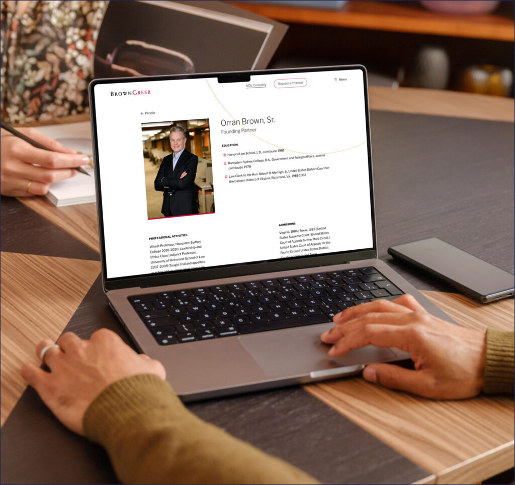 A laptop is open on a wooden table, displaying a professional profile for "Orran Brown, Sr.," who is identified as a founding partner. The screen shows sections for education, professional activities, and admissions, alongside a photo of the individual. A hand rests on the laptop's trackpad, while another hand holds a pen, indicating someone is taking notes on a notepad nearby. A smartphone is placed next to the laptop on the table. The setting appears to be a collaborative workspace.