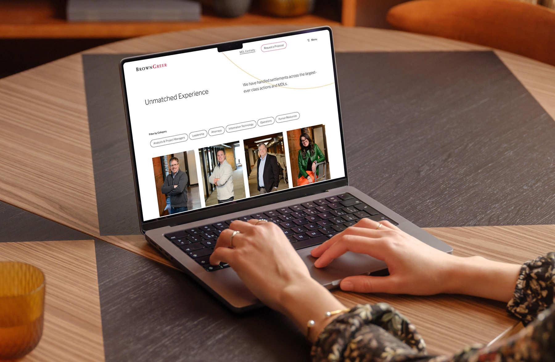 A laptop is open on a wooden table, displaying a webpage titled "Unmatched Experience" from BrownGreer. The screen features a grid of images showcasing several individuals, along with filter options for categories like "Leadership" and "Information Technology." A person's hands are visible on the keyboard, and a textured glass cup is placed nearby. The table has a mix of light and dark wood patterns.