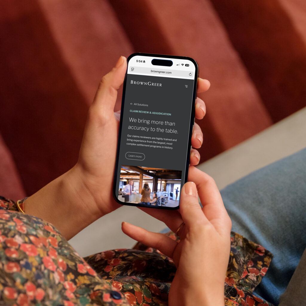 A person is holding a smartphone displaying a webpage for BrownGreer, featuring a dark background with white text. The section highlighted is titled "Claim Review & Adjudication," emphasizing the expertise of claims reviewers. The phone is positioned in front of the person's floral-patterned clothing, with a soft, textured surface visible in the background. The setting appears cozy, with warm tones in the surrounding environment.