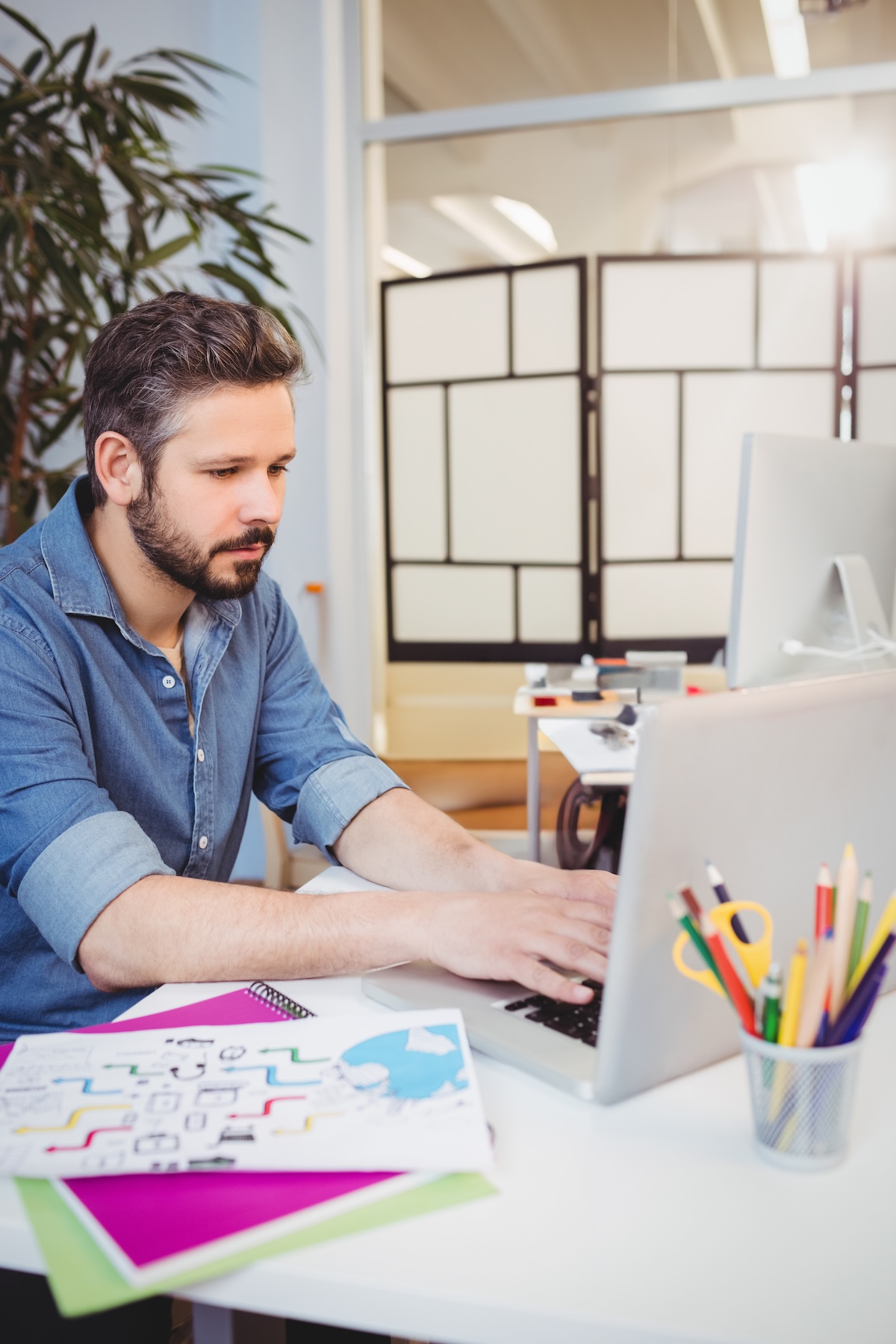 A man with short dark hair and a beard, wearing a blue denim shirt with rolled-up sleeves, is sitting at a white desk working on a laptop. On the desk, there are colorful folders, a sheet with diagrams or drawings, and a metal cup holding various colored pencils and scissors. In the background, there is a large plant and a room divider with a geometric pattern.