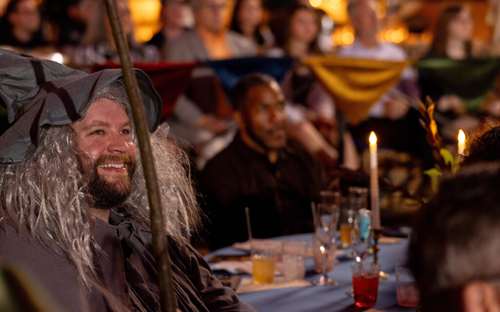 A man dressed as a wizard with a gray wig and a large pointed hat is smiling. He is seated at a table with drinks and lit candles, and there are colorful triangular flags hanging in the background. Other people are seated around the table, but they are out of focus.