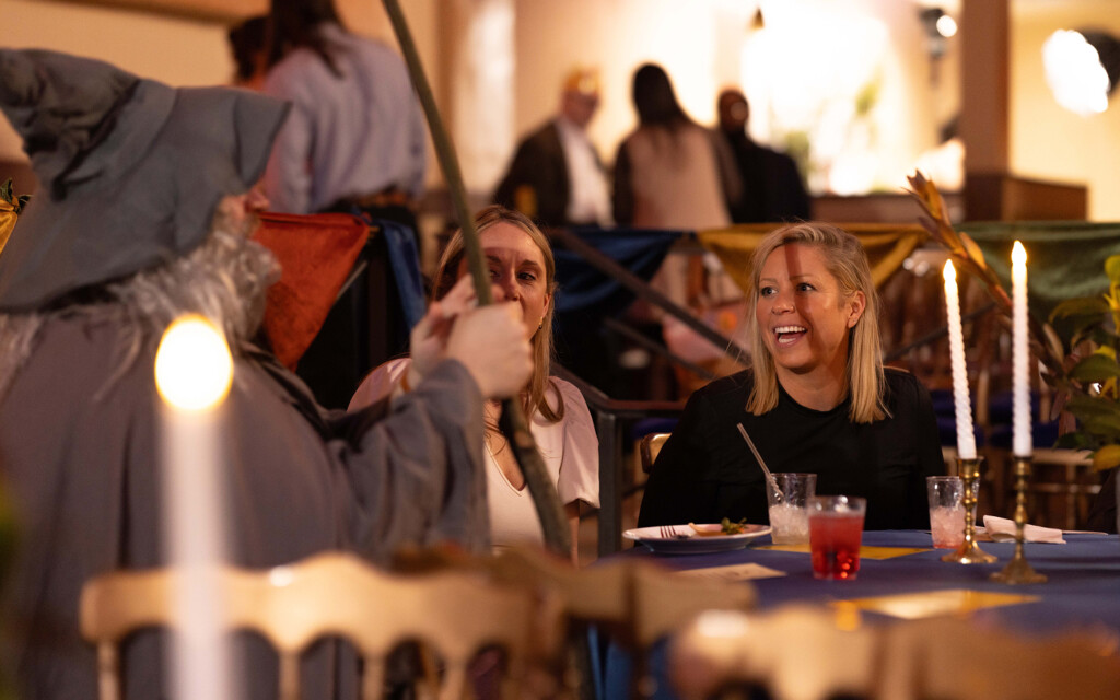 Two women sitting at a table with drinks and plates, engaged in conversation with a person dressed as a wizard holding a staff. The table is covered with a blue tablecloth and decorated with two lit candles in brass candle holders. The background shows blurred figures and colorful banners.