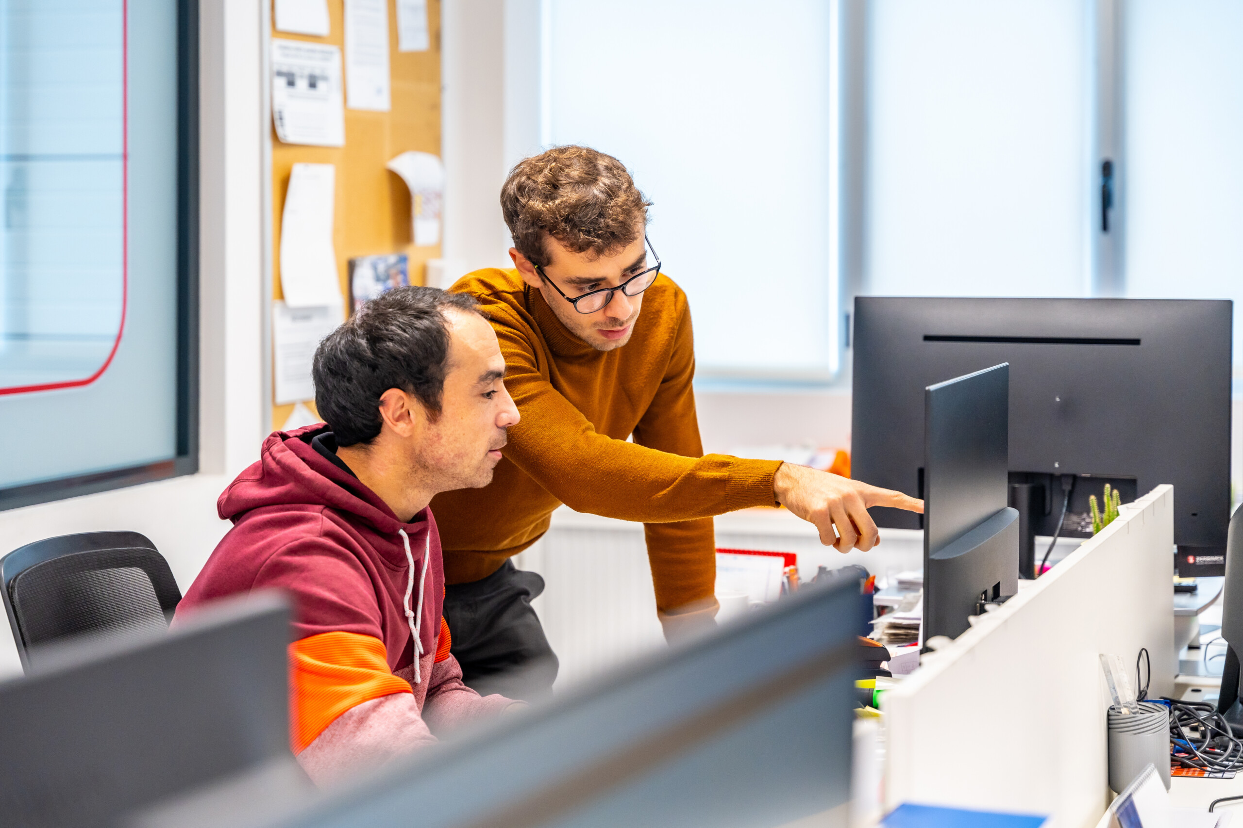 Two men are working together at a desk in an office. One man, wearing a maroon and orange hoodie, is seated and looking at a computer screen. The other man, wearing glasses and a brown sweater, is standing and pointing at the computer screen. The workspace includes multiple monitors, office supplies, and a bulletin board with papers pinned on it in the background.