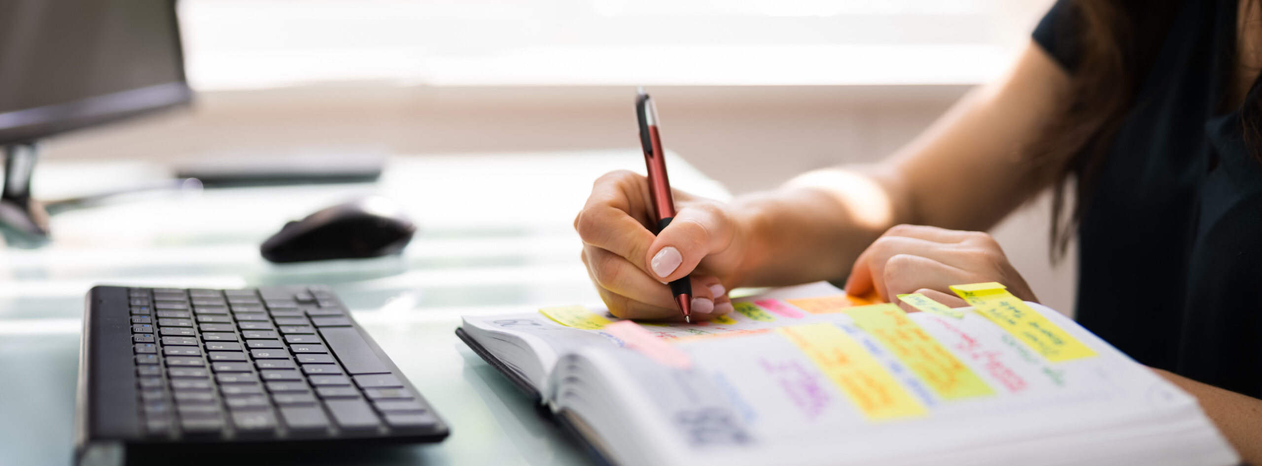 A hand with light pink nail polish holding a red and black pen, writing in an open planner or notebook filled with colorful sticky notes and handwritten notes. The scene is set on a glass surface with a keyboard partially visible on the left side.