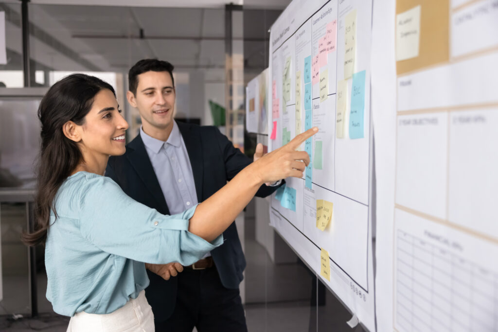 A woman and a man are standing in an office, looking at a large board covered with charts and colorful sticky notes. The woman, wearing a light blue blouse, is pointing at the board while smiling, and the man, dressed in a dark suit with a light blue shirt, is attentively observing the board. The setting appears to be a collaborative work environment.
