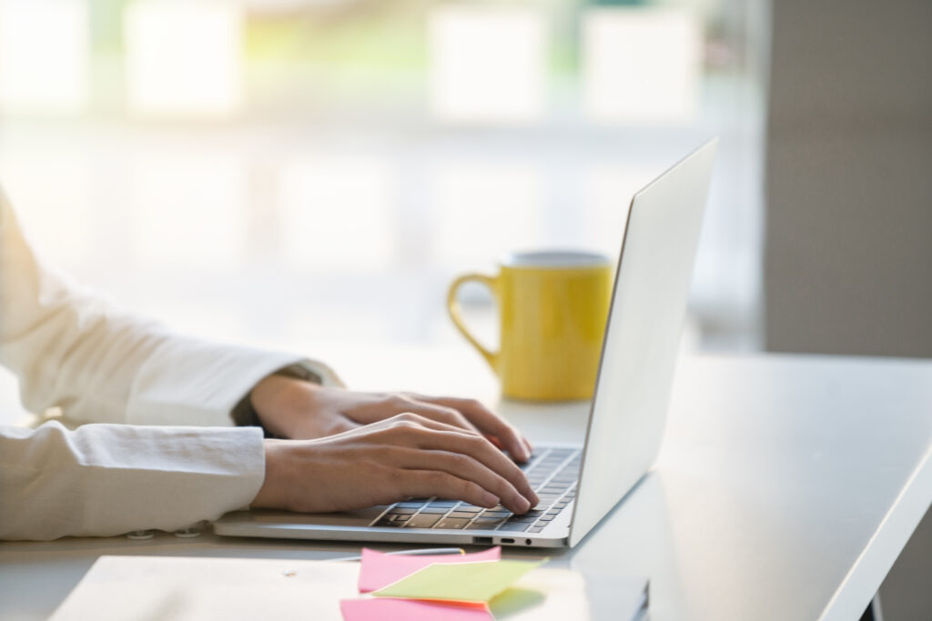 Hands typing on a laptop keyboard on a white desk with a yellow mug and colorful sticky notes nearby.