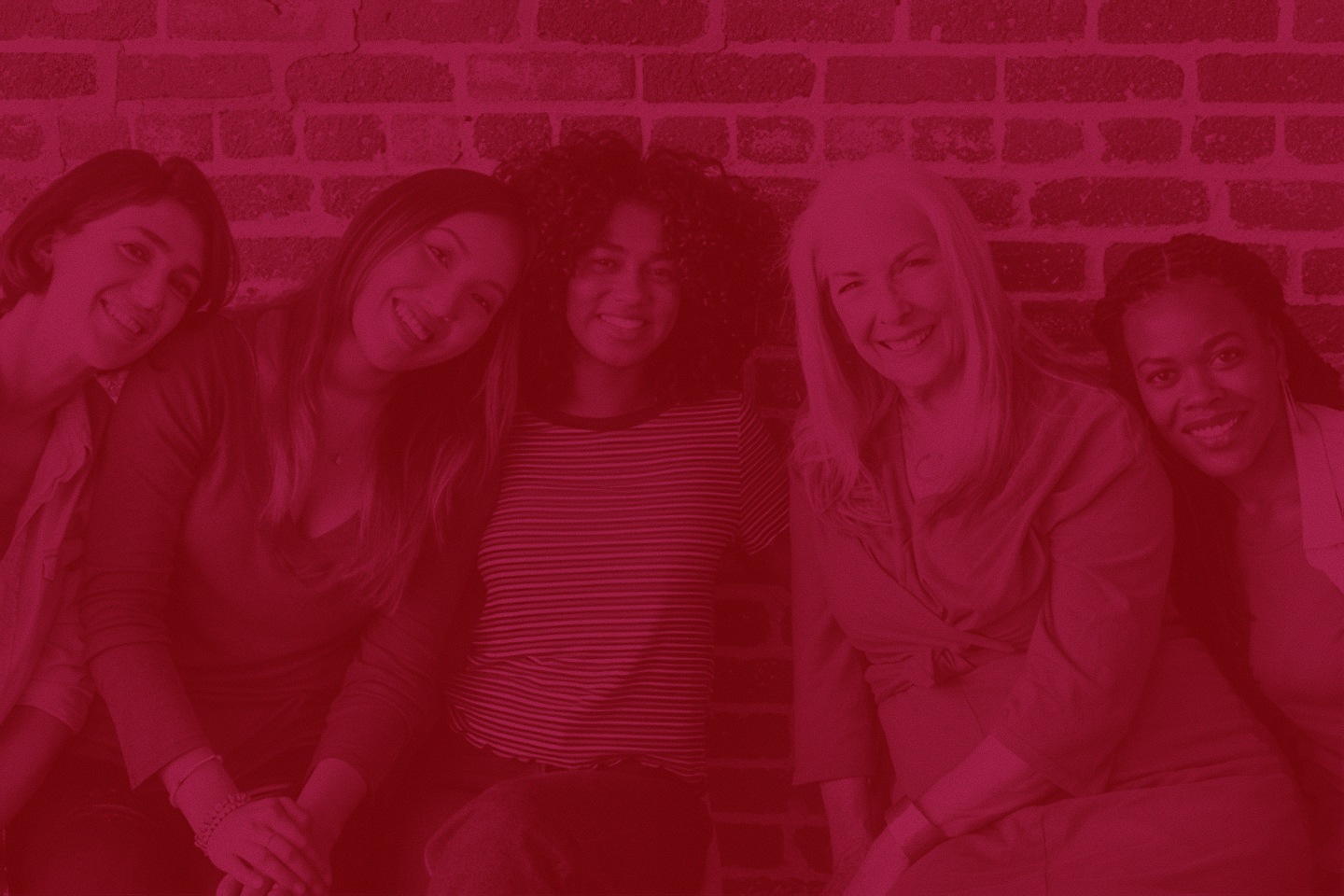 Five diverse women sitting close together in front of a brick wall, smiling warmly at the camera.