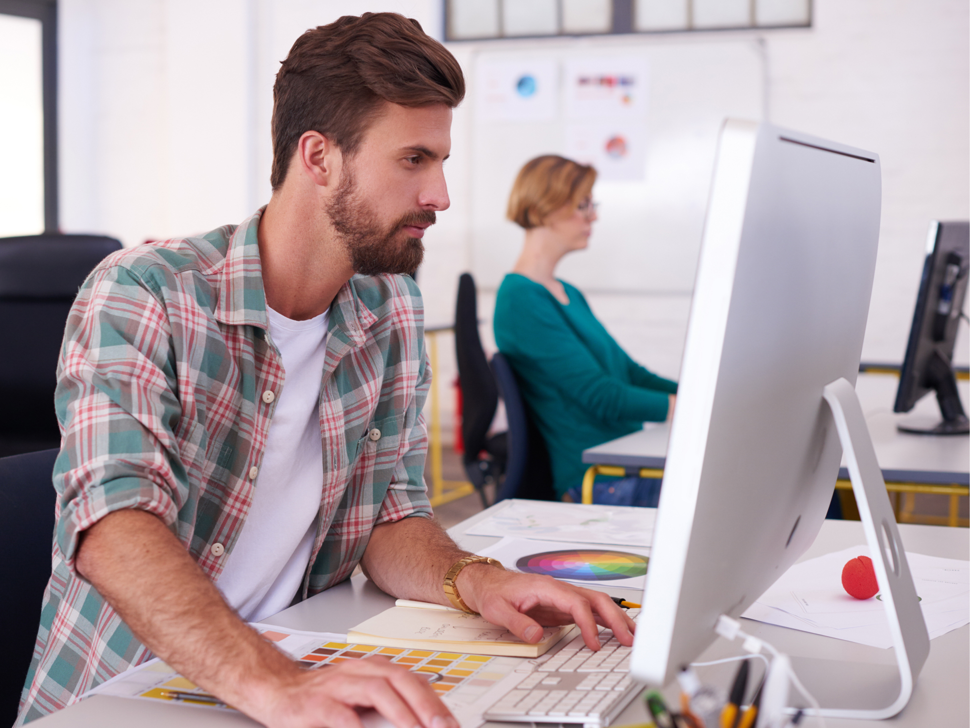 Man using a desktop computer at a desk in an office, with a woman working at another computer in the background.