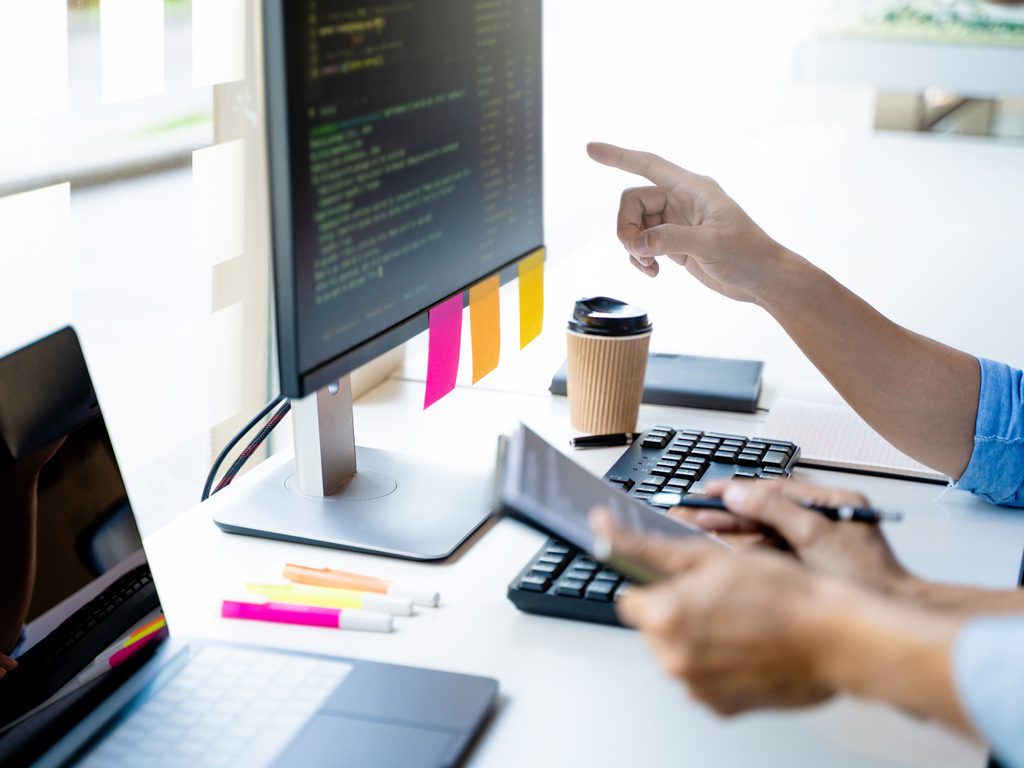 Person pointing at a computer monitor with code, beside sticky notes, a coffee cup, keyboard, and tablet on a desk.