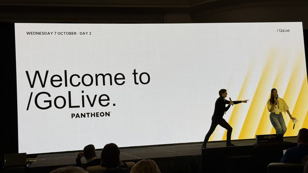 Conference stage with large screen reading “Welcome to /GoLive. PANTHEON” and “WEDNESDAY 7 OCTOBER · DAY 2,” with two speakers onstage