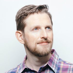Portrait of a man with short hair and a beard wearing a purple plaid shirt, looking to the side.